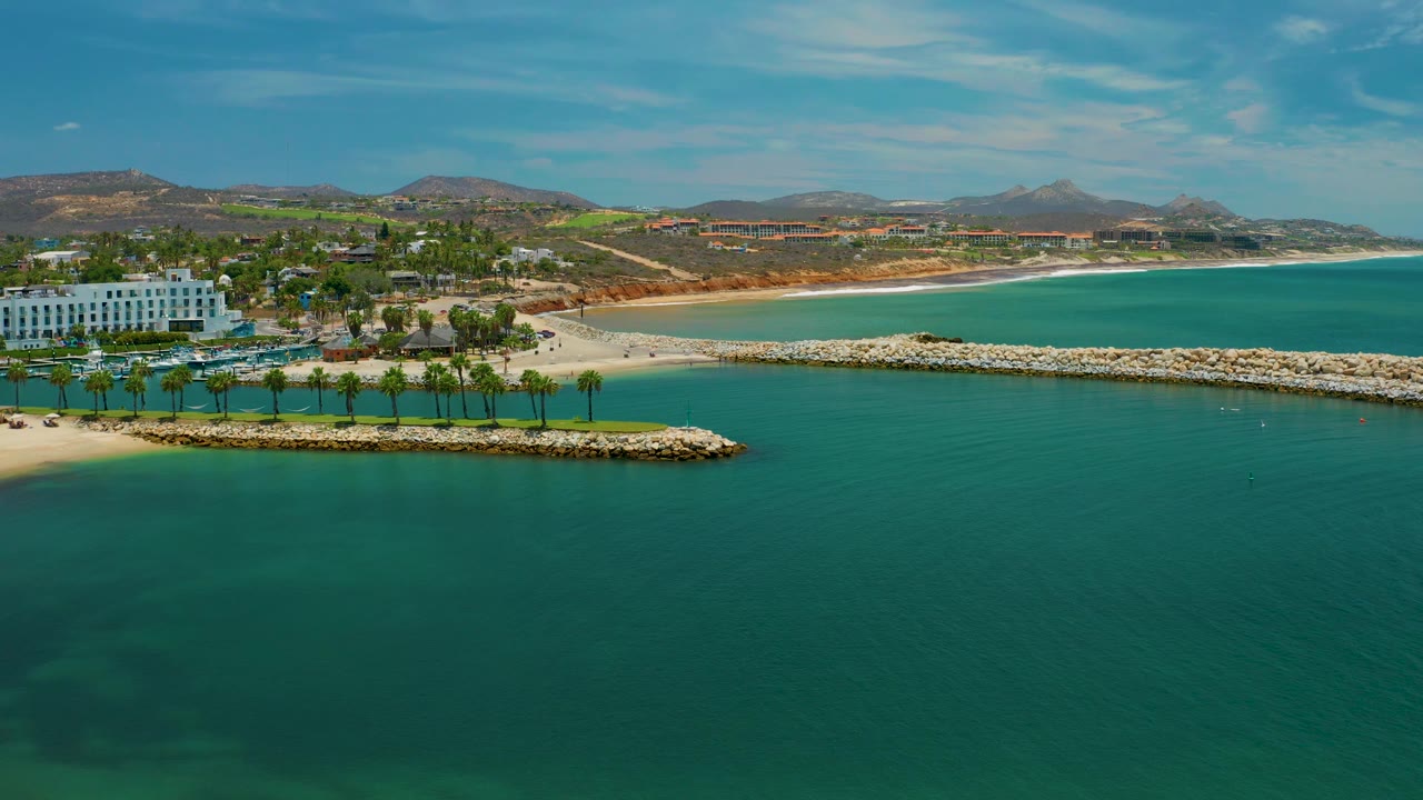 Wide San Jose Del Cabo beach frame with a larger area of open turquoise bay to the right, sand curving along the shore, palms and a white beachfront building at left, and no visible people.