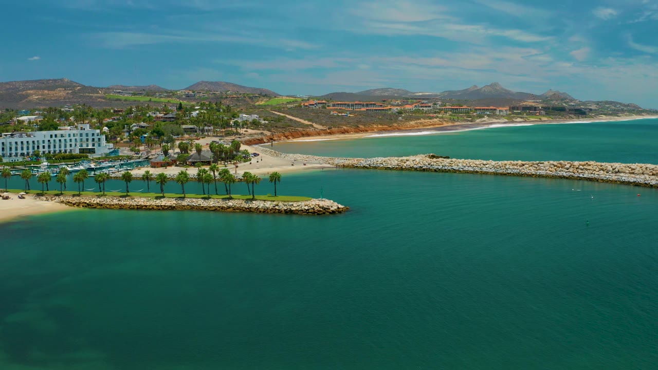 Elevated San Jose Del Cabo coastal frame showing the palm row and breakwater more centrally, with a white beachfront building on the left, turquoise water below, hills beyond, and no visible people.
