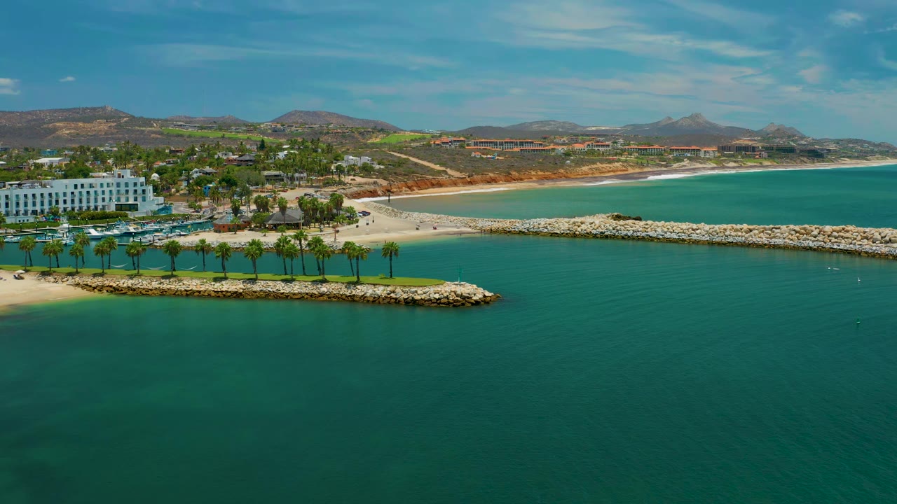 San Jose Del Cabo coastline seen from above with teal water filling the foreground, pale rock barriers crossing the frame, a palm-lined beach zone, white buildings on the left, and no visible people.