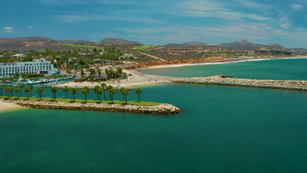 Aerial frame over San Jose Del Cabo with turquoise sea in front, a rock breakwater with green palms, white resort buildings at left, beach and dry hills beyond, and no visible people.