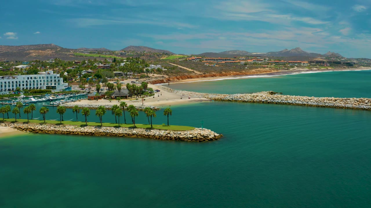 Aerial frame over San Jose Del Cabo with the palm-lined rock barrier low in the image, white buildings and landscaped grounds at left, a sandy beach curving across the center, and no visible people.