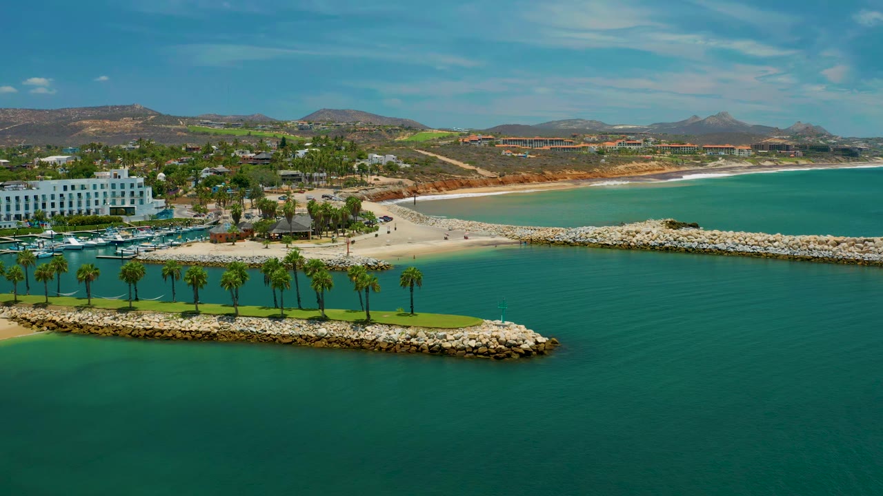 Aerial frame from San Jose del Cabo, Mexico, showing the white building farther left, calm lagoon water below, a row of palms on the breakwater, sandy beach in back, and no visible people.