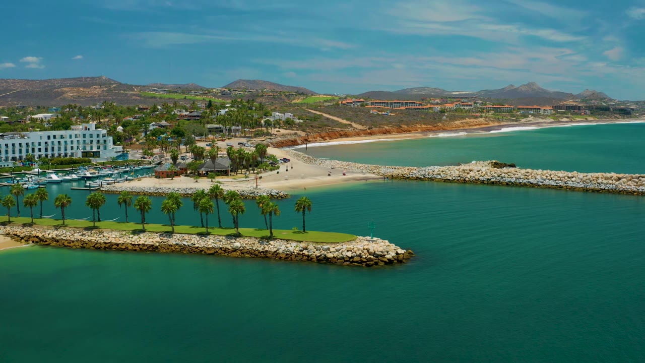San Jose del Cabo aerial view with the white seaside building near the left edge, green palm strip across the breakwater, bright turquoise lagoon water, beach behind it, and no visible people.