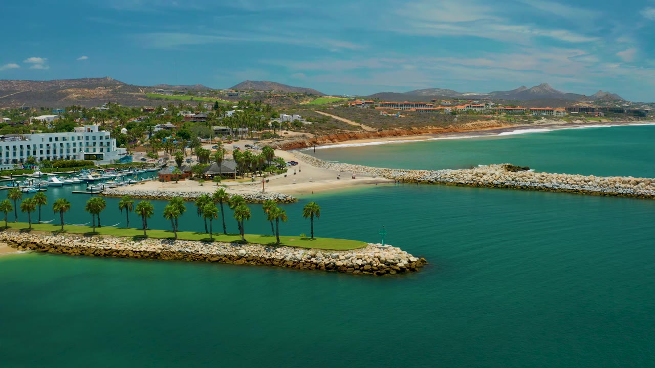 Elevated San Jose del Cabo frame with deeper green water in the foreground, a palm row on the rock breakwater, the white building left of center, a pale beach curve, and no visible people.