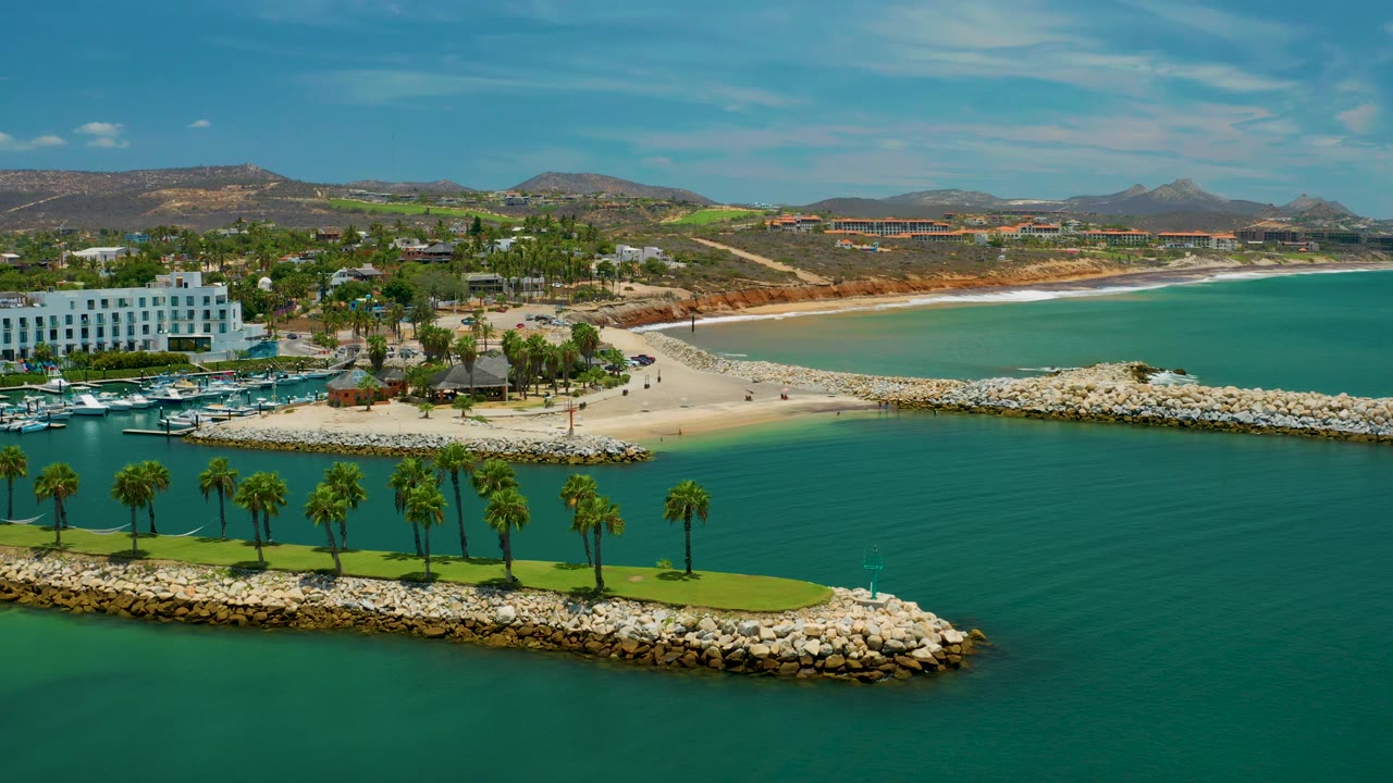 Aerial view in San Jose Del Cabo showing the palm breakwater across the lower frame, dark green inlet water, pale beach, marina boats near a white hotel, distant hills, and no visible people.
