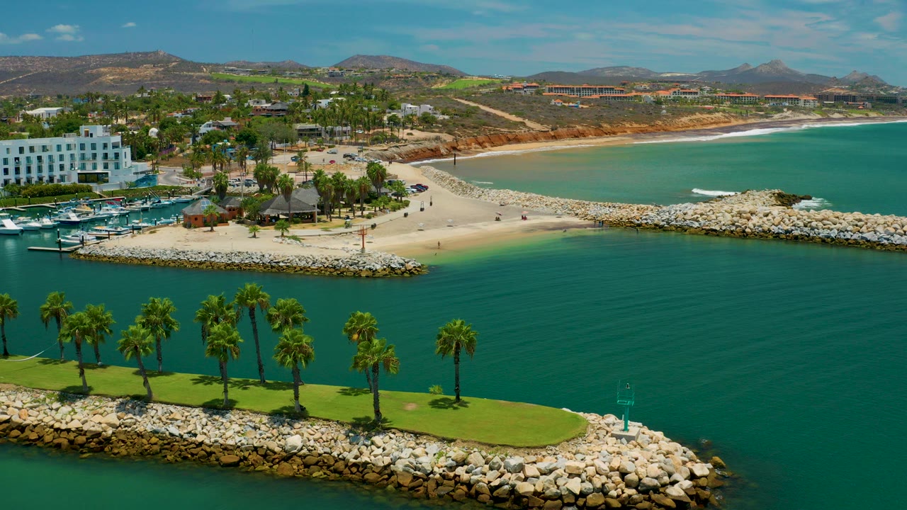 Aerial frame in San Jose del Cabo showing turquoise water in front, a palm-lined lawn on a rock edge, marina boats, white buildings, sandy beach, low hills, and no visible people.