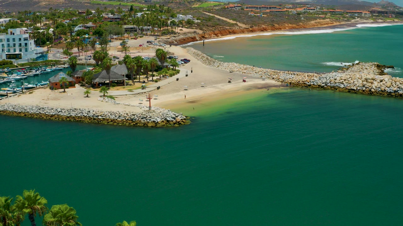 Aerial view in San Jose Del Cabo, Mexico showing turquoise water in the foreground, a sandy beach and rock breakwater, palms, marina boats, low buildings, distant hills, and tiny people on shore.