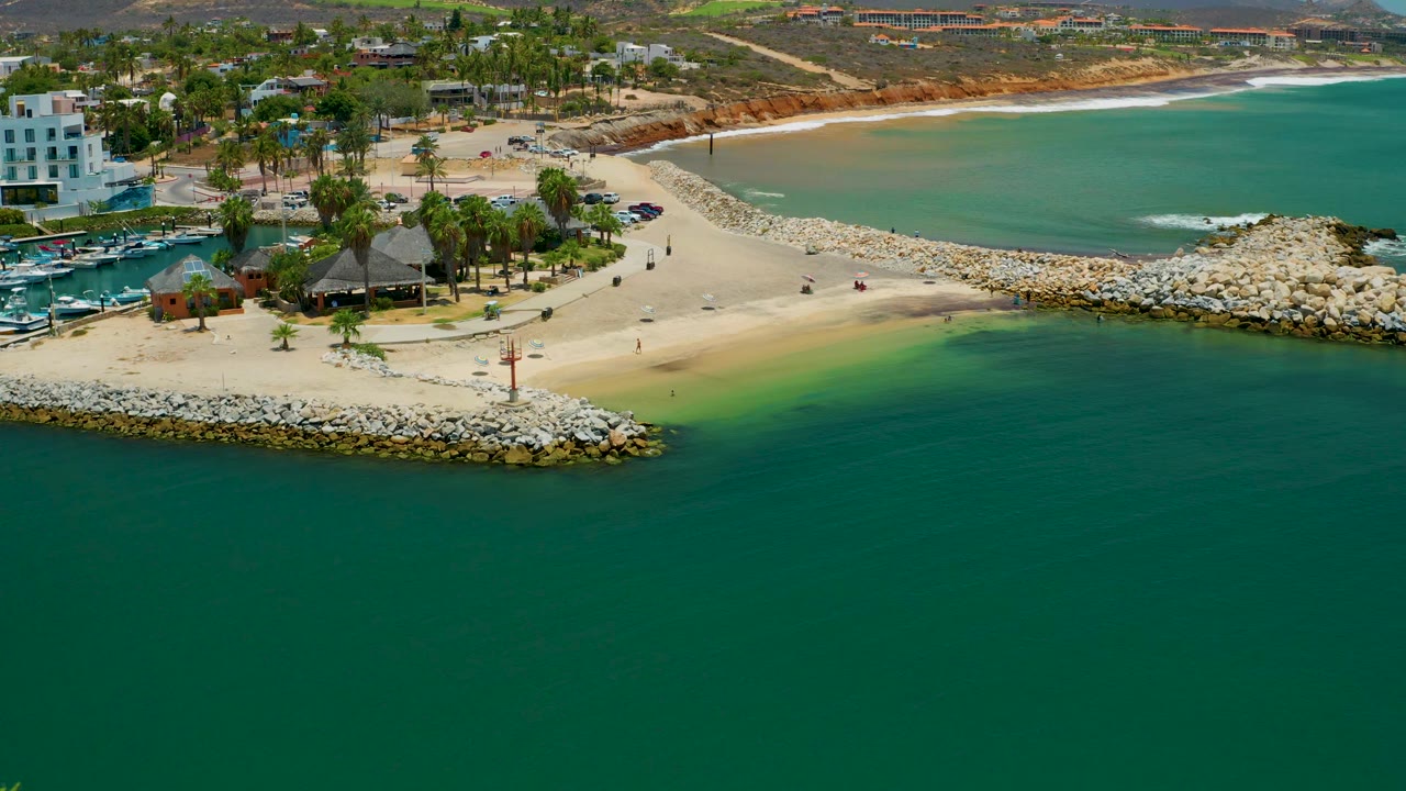 Wide aerial view in San Jose Del Cabo, Mexico with turquoise shallows near the sand, a rocky point at the breakwater, palm-lined park areas, shoreline buildings, hills, and tiny figures on the beach.