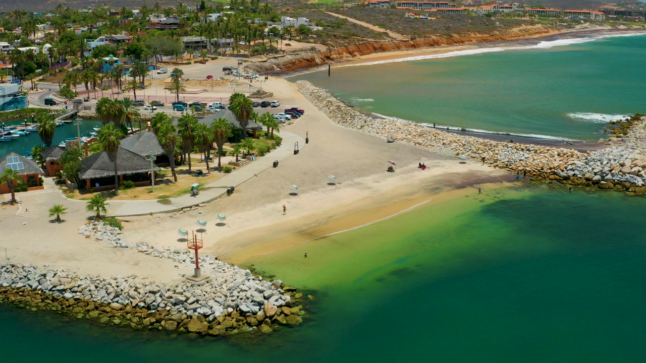 High aerial view of a San Jose del Cabo beach with a rock jetty in the lower foreground, palms and thatched roofs at left, and blue-green bay water at right.