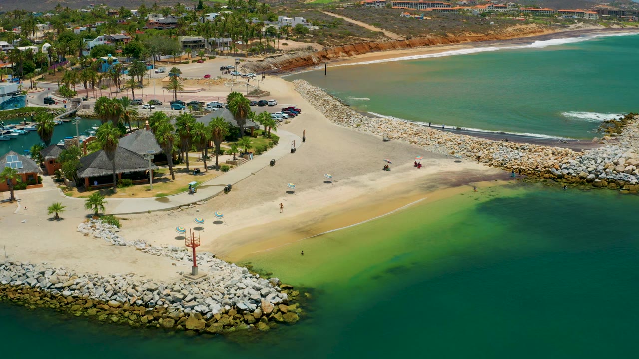 Aerial frame over San Jose del Cabo shoreline with the curved rock breakwater crossing the beach, green shallows in the lower right, and palm landscaping beside parked cars.