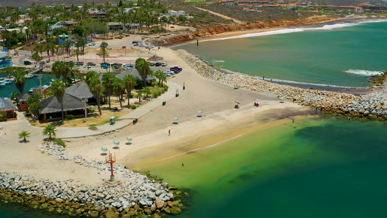 San Jose del Cabo beach seen from above with green shallows in the foreground, a long stone jetty leading toward the bay, and a pale surf line along the far shore.