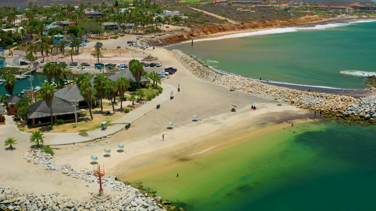 High view of the San Jose del Cabo beach area with thatched roofs and palms at lower left, open sand through the center, and the rock breakwater separating beach from bay.