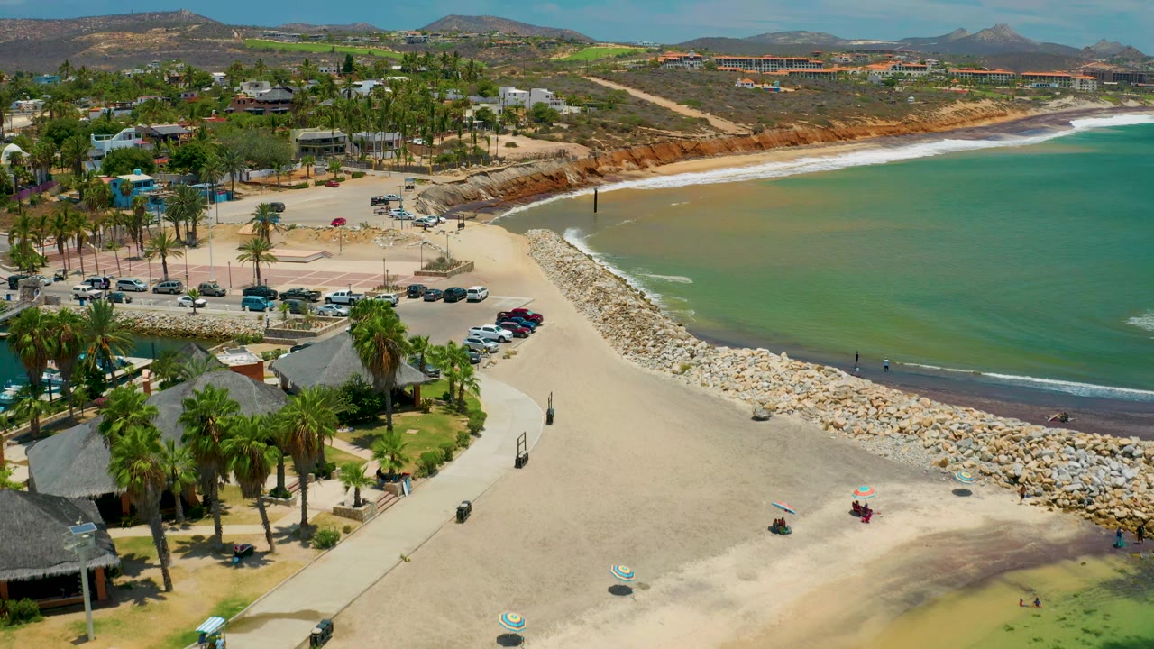 San Jose Del Cabo aerial view with palms and parked cars in the foreground, a pale sand beach and rock breakwater in the middle, green-blue water to the right, and no visible people.