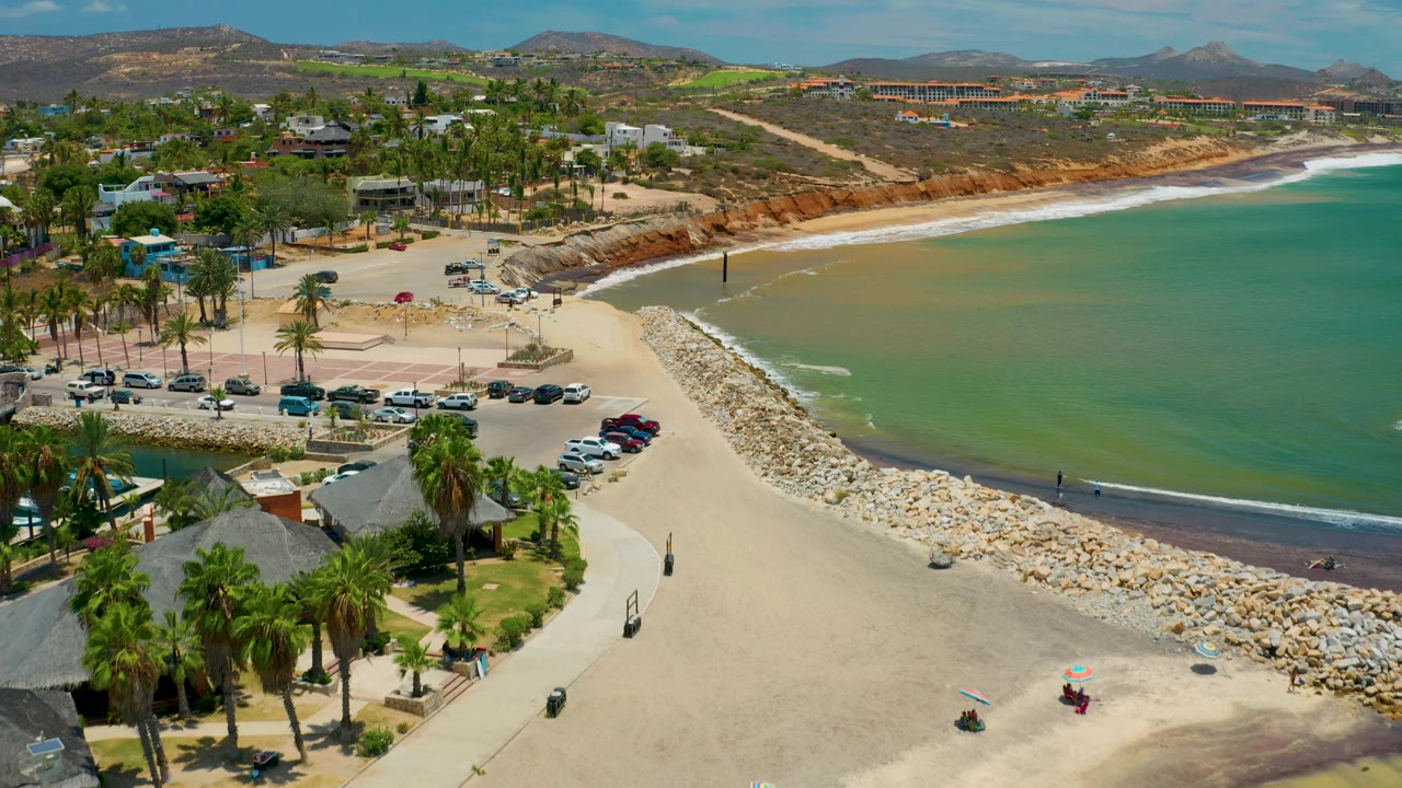 Aerial frame in San Jose Del Cabo showing beach access, rows of parked cars, palm trees, a curved shoreline with small surf, hillside buildings, and no visible people.