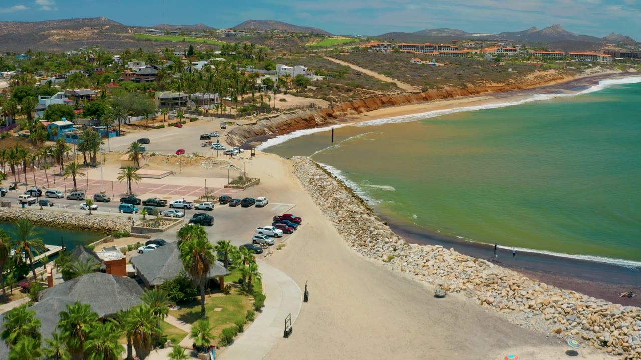 Wide aerial of beach, breakwater, parking area, palms, green water, and hills.