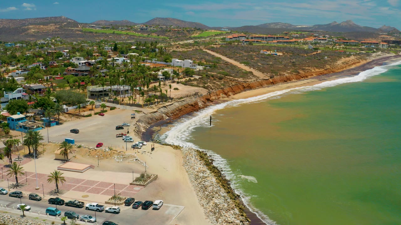 Aerial frame from San Jose Del Cabo, Mexico showing green sea water meeting a curved beach, a rock edge, palm trees, parked cars, low coastal buildings, distant hills, and no visible people.