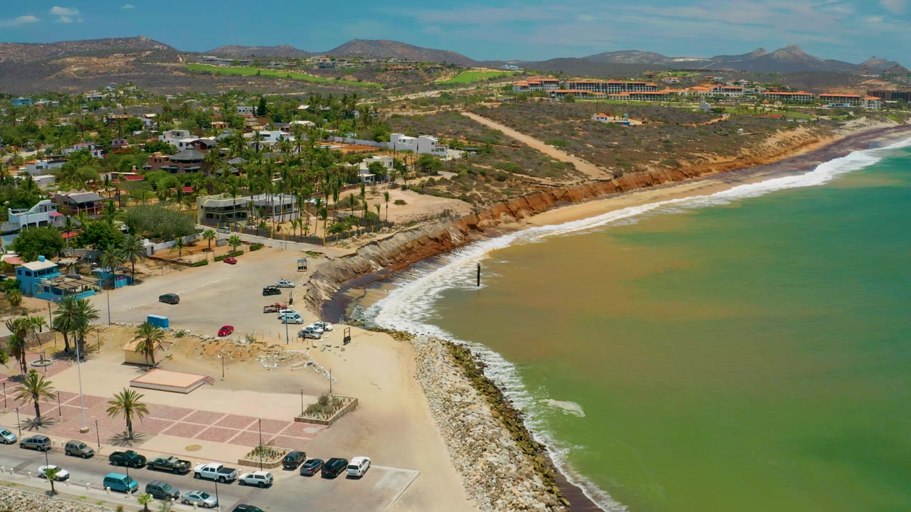 Second aerial frame of the San Jose Del Cabo coast with the white wave line slightly farther along the curved shore, green water to the right, parked cars below, hillside buildings beyond, and no visible people.