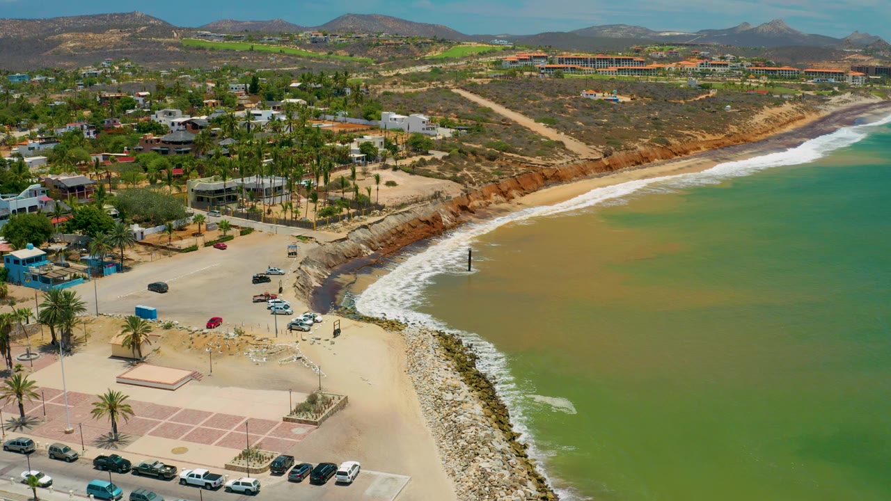 Aerial view over San Jose Del Cabo with more open green water filling the right side, white surf tracing the shore, a small parking area and palms at lower left, resort-area buildings in back, and no visible people.