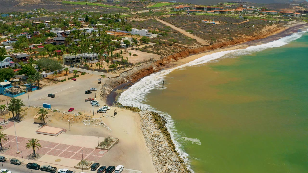 Drone-like view of a beach cove with green sea, white waves, palms, cars, and coastal buildings.