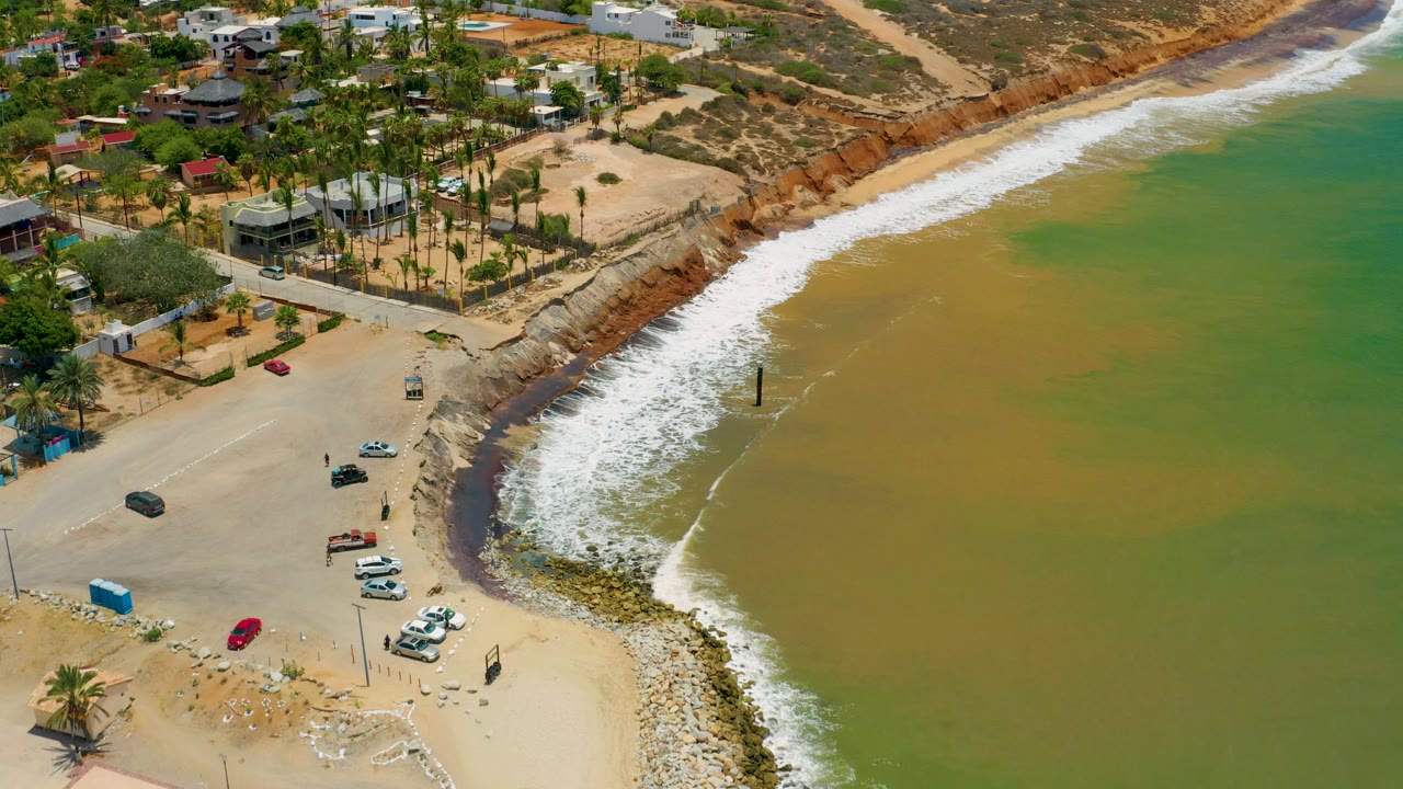 Aerial frame of San Jose Del Cabo beach access with white surf curving along green-brown water, parked cars on sand, palm trees, homes, and tiny visible people near the shore.