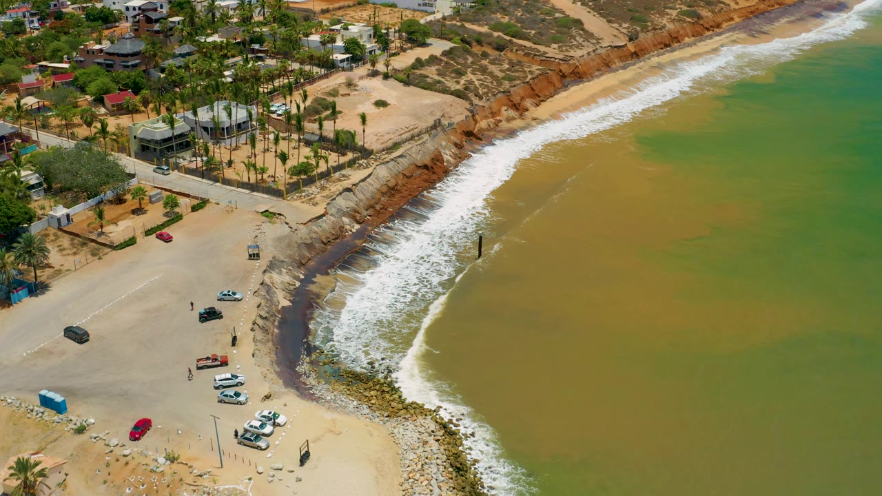 Aerial San Jose Del Cabo frame with the camera view still angled over parked cars, a broad sweep of foamy surf, reddish shoreline rock, palm-lined homes, and tiny visible people on the sand.