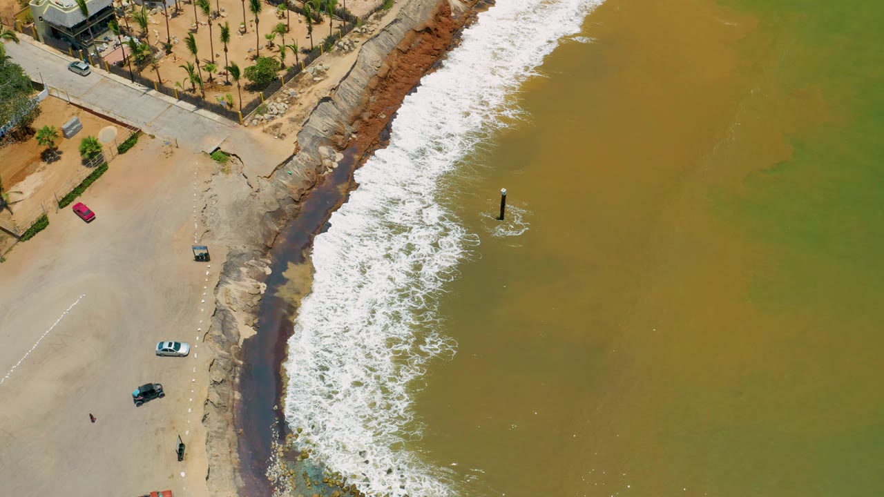 Top-down aerial view in San Jose Del Cabo, Mexico, showing a diagonal shoreline with white surf, murky green-brown water, sandy open beach, road, palms, parked cars, and no visible people.