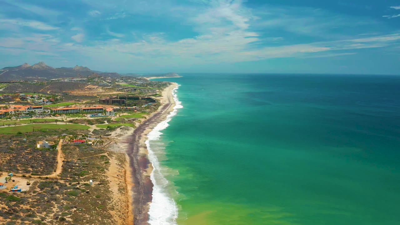 Wide aerial frame over San Jose Del Cabo with blue-green ocean, white breakers meeting the beach, coastal buildings to the left, mountains beyond, and no visible people.