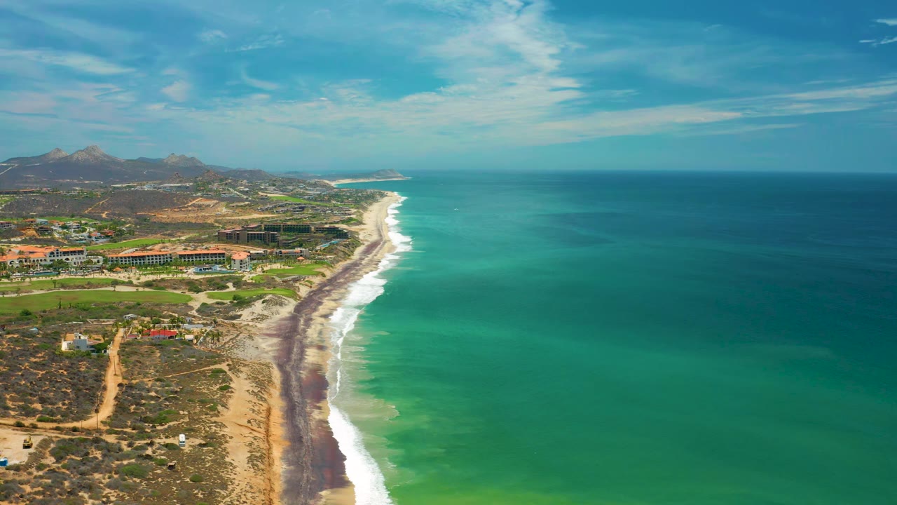 At 0:01, the San Jose del Cabo shoreline runs diagonally between green-blue ocean and dry inland terrain, with resort-scale buildings, roads, hills, sky, and no visible people.