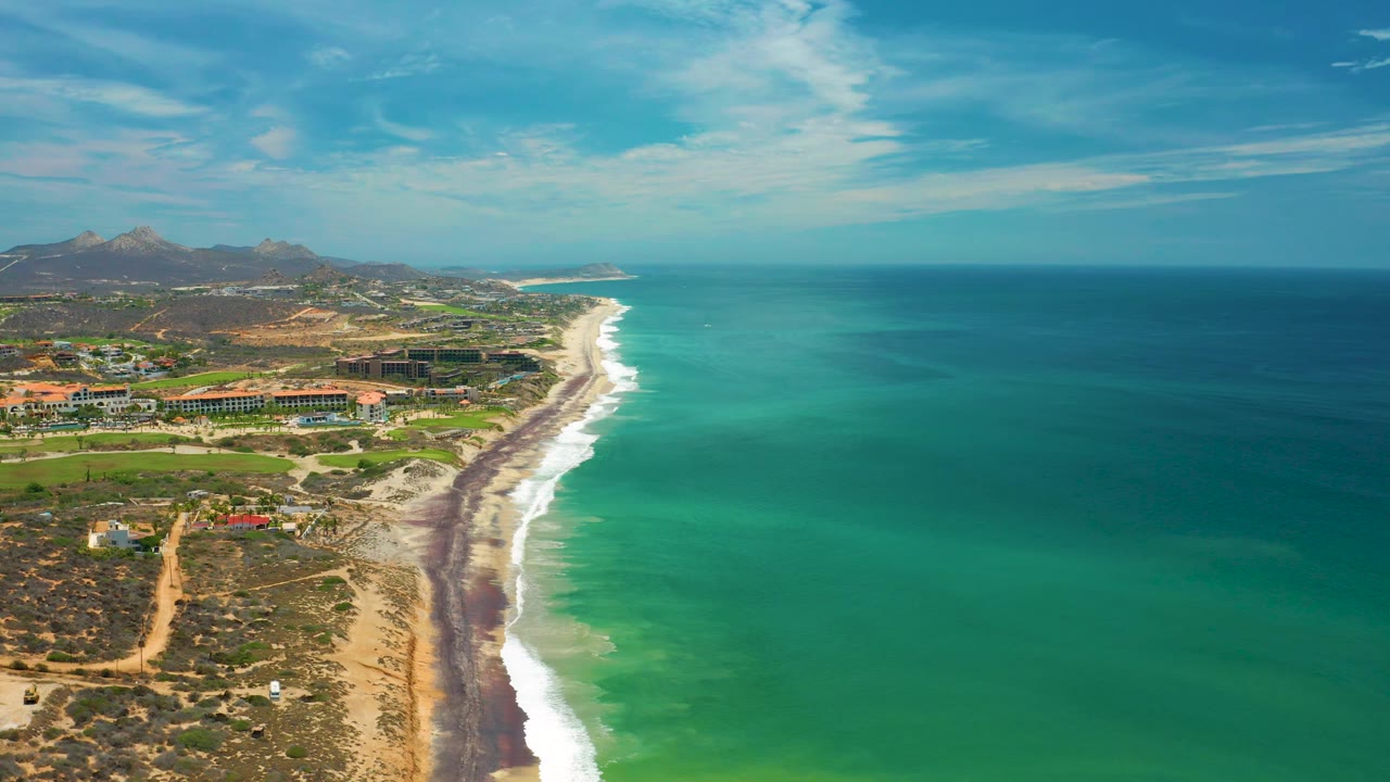 At 0:02, a wide aerial composition over San Jose del Cabo shows more open ocean across the right side, a bright surf edge, inland roads and buildings, distant mountains, and no visible people.