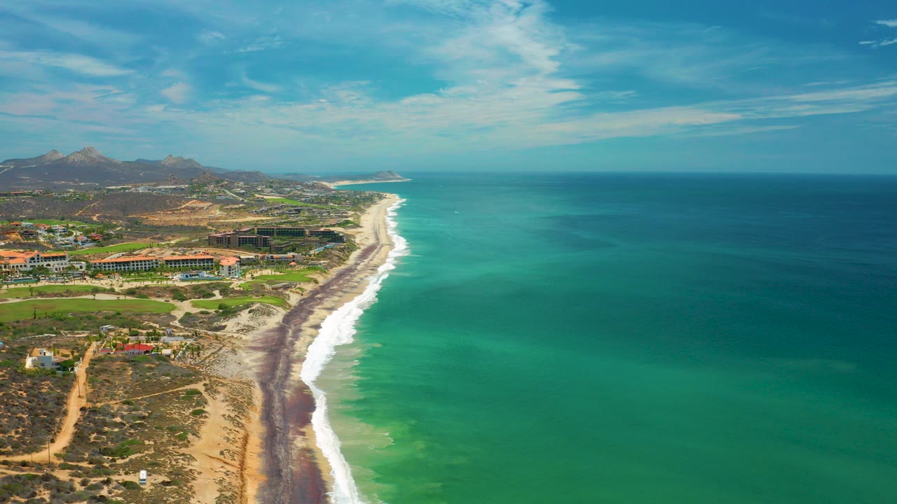 Aerial view over San Jose del Cabo, Mexico, with turquoise water on the right, a sandy surf line on the left, distant hills, and no visible people.