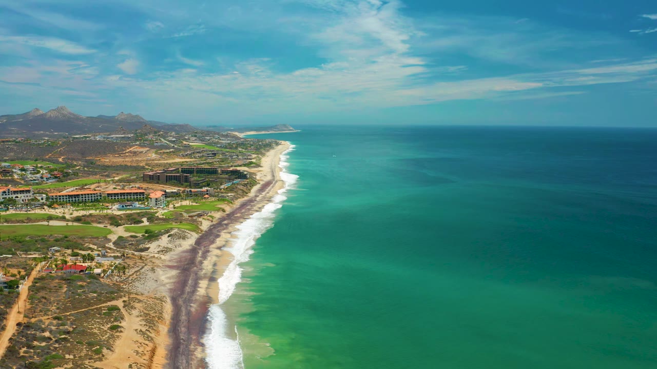Aerial frame from San Jose Del Cabo, Mexico, with the shoreline running diagonally through the image, blue-green water filling the right side, resort buildings inland, hills beyond, and no visible people.