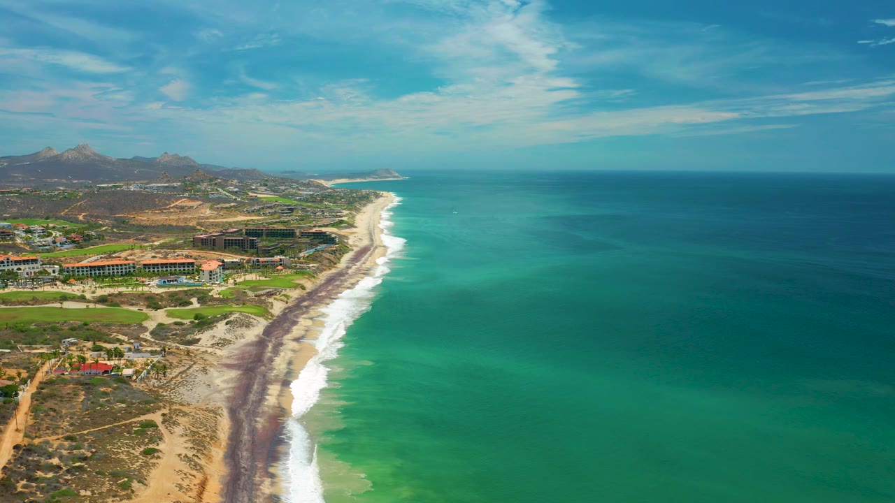Aerial view of the San Jose Del Cabo coastline with broad open ocean on the right, a narrow sandy beach and surf strip, inland resort structures, hazy hills in the distance, and no visible people.