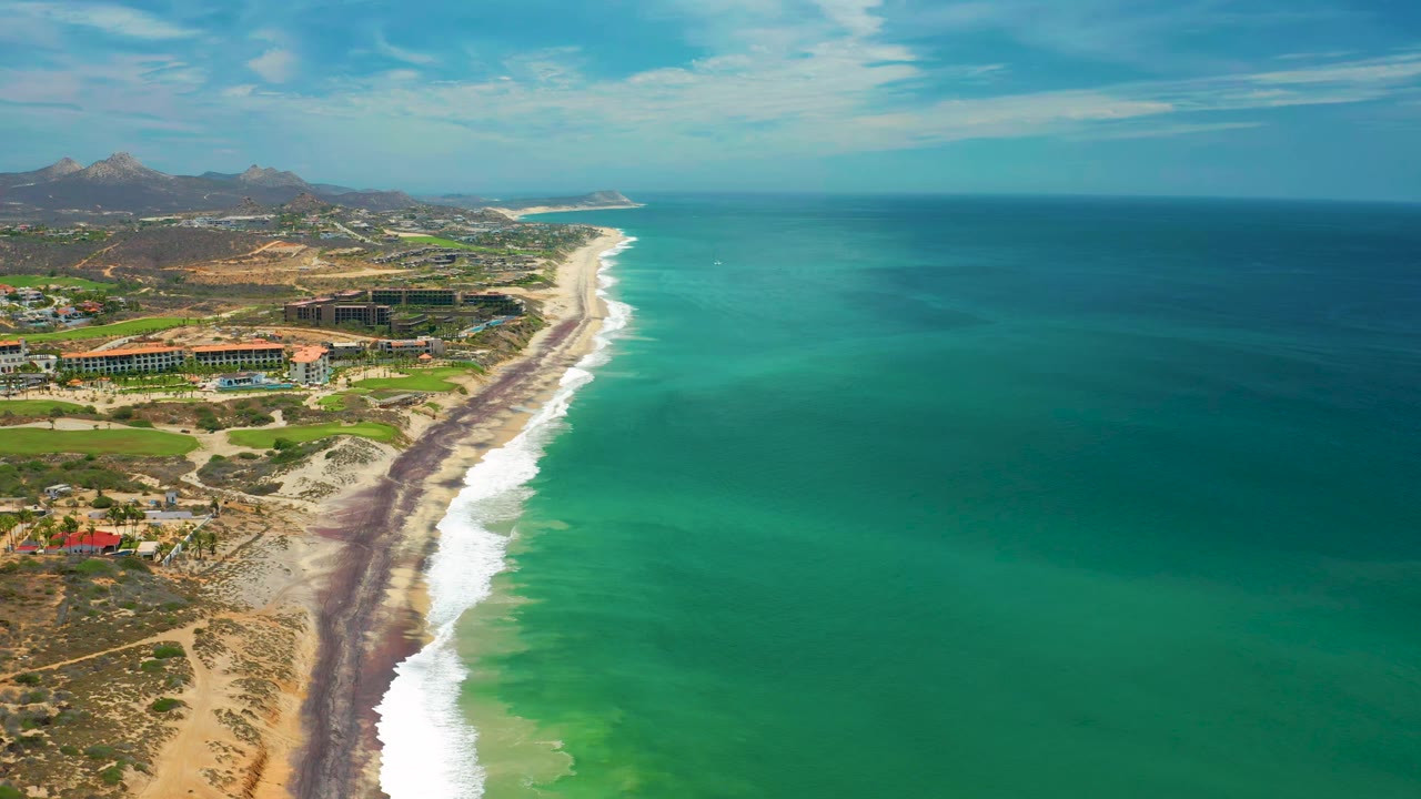 High aerial frame over San Jose del Cabo with the surf line curving along the beach, green-blue ocean filling the right side, low buildings and homes inland, and no visible people.