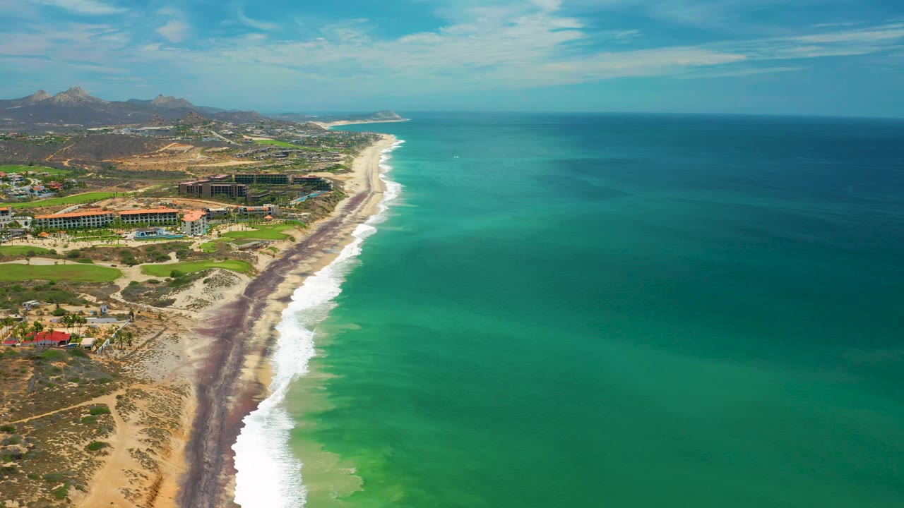 Drone-style view of the San Jose del Cabo shoreline with a dark band along the sand, white waves breaking near shore, broad green-blue water, inland buildings, distant hills, and no visible people.