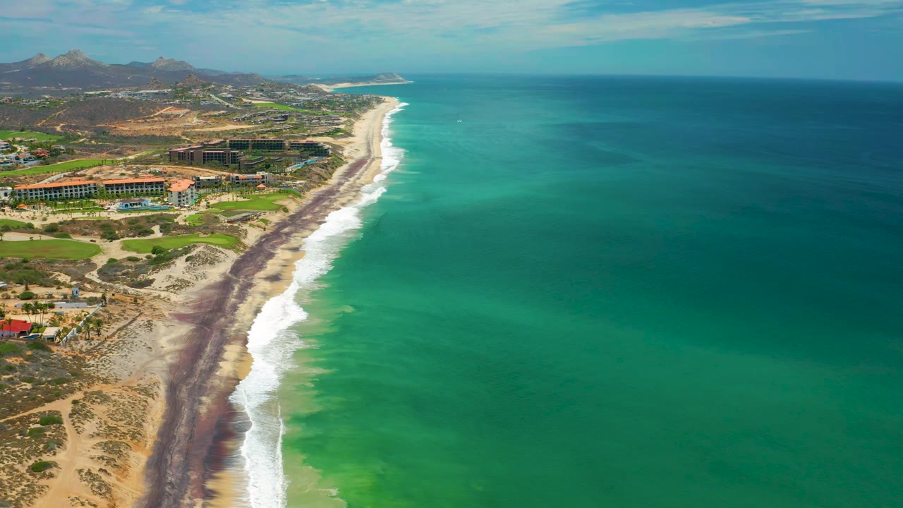 Aerial view in San Jose del Cabo looking along the beach as white waves meet a dark shoreline band, with resort grounds inland and no visible people.