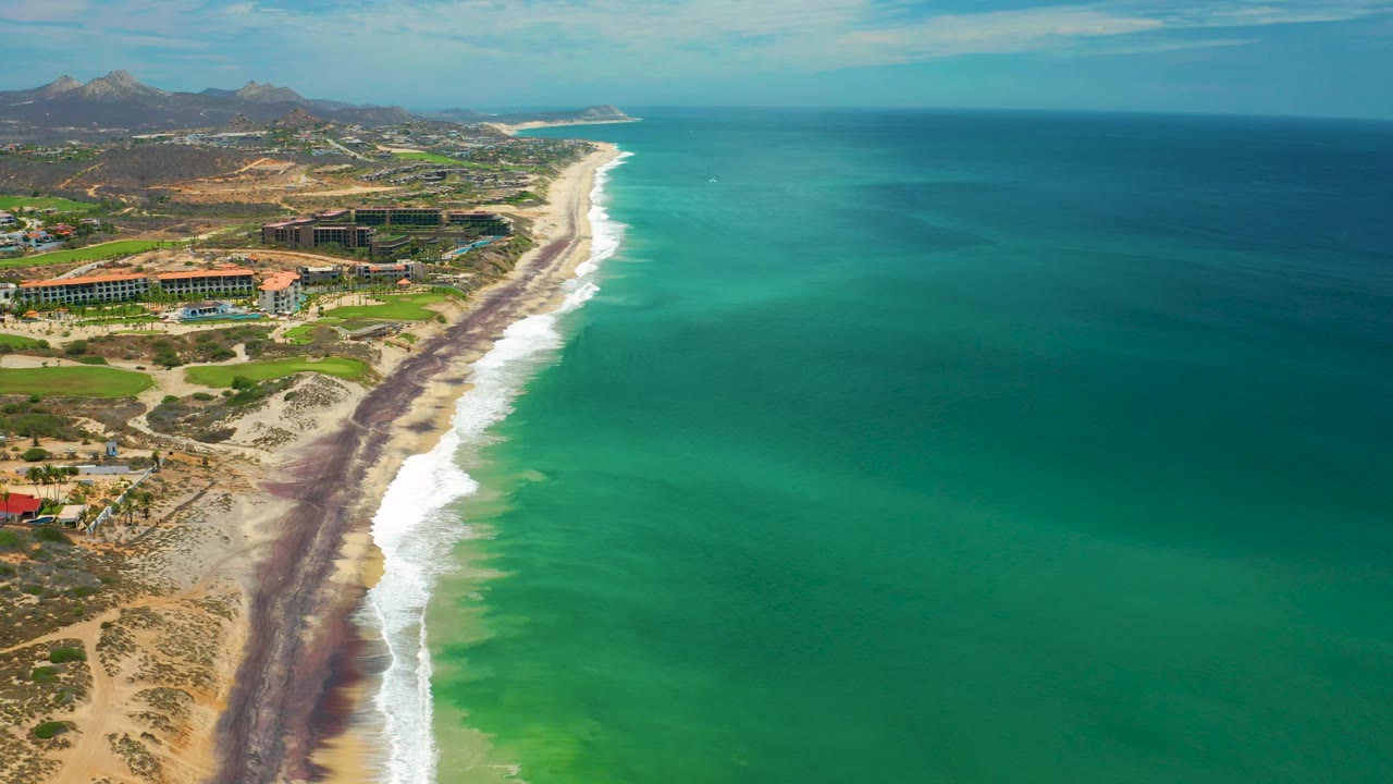 High aerial frame of the San Jose del Cabo coast with open blue-green water filling the right side, resort buildings near the beach, distant hills, and no visible people.