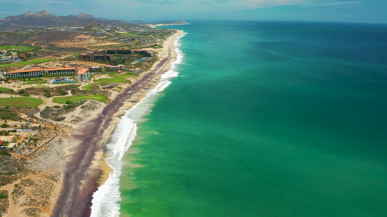 Aerial view of San Jose Del Cabo coastline at the start of the clip, with green-blue water on the right, surf along a dark tide line, coastal buildings on the left, distant hills, and no visible people.