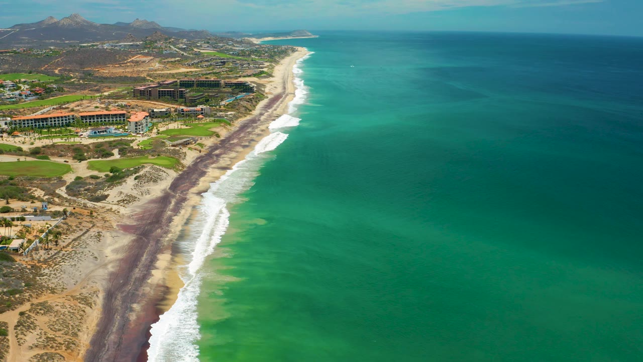 Oblique aerial view over San Jose Del Cabo beach, with a long surf line, emerald water extending to the horizon, inland buildings and roads at left, hills in the background, and no visible people.