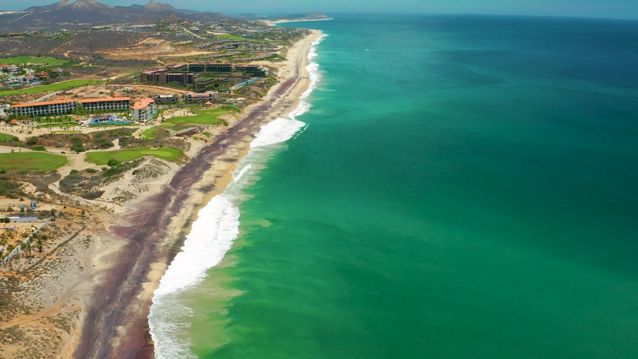 High aerial view over San Jose del Cabo, Mexico, showing turquoise water beside a long surf line, sandy beach, resort buildings, green landscaping, inland hills, and no visible people.