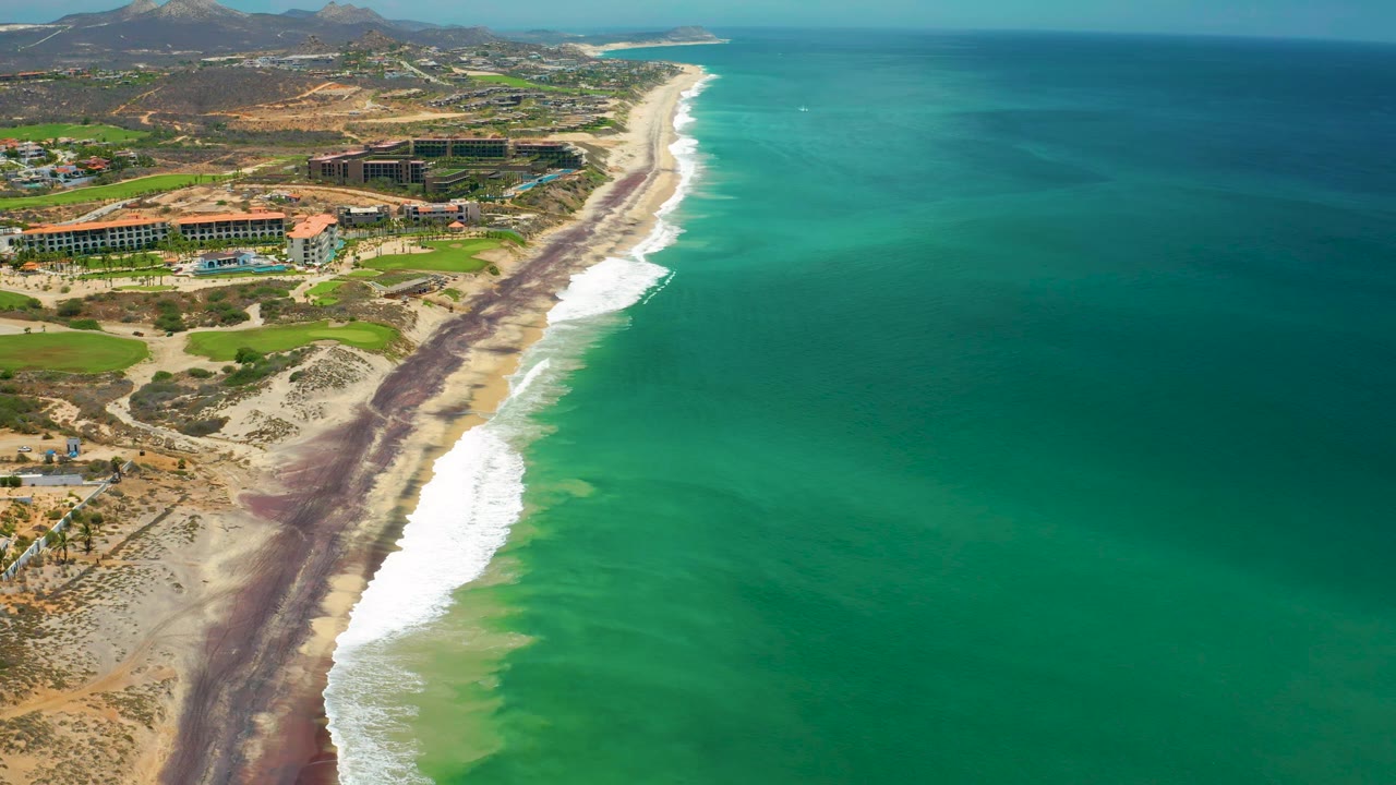 Aerial frame in San Jose del Cabo, Mexico, with more open green-blue ocean filling the right side, white waves on the beach, resort roofs inland, and no visible people.