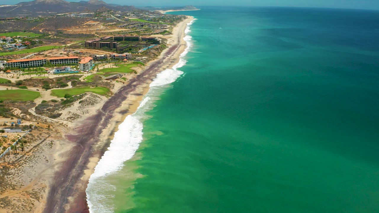High coastal view over San Jose del Cabo, Mexico, where the beach and surf curve toward the distance, open sea dominates the right half, inland hills sit beyond the resort area, and no visible people appear.