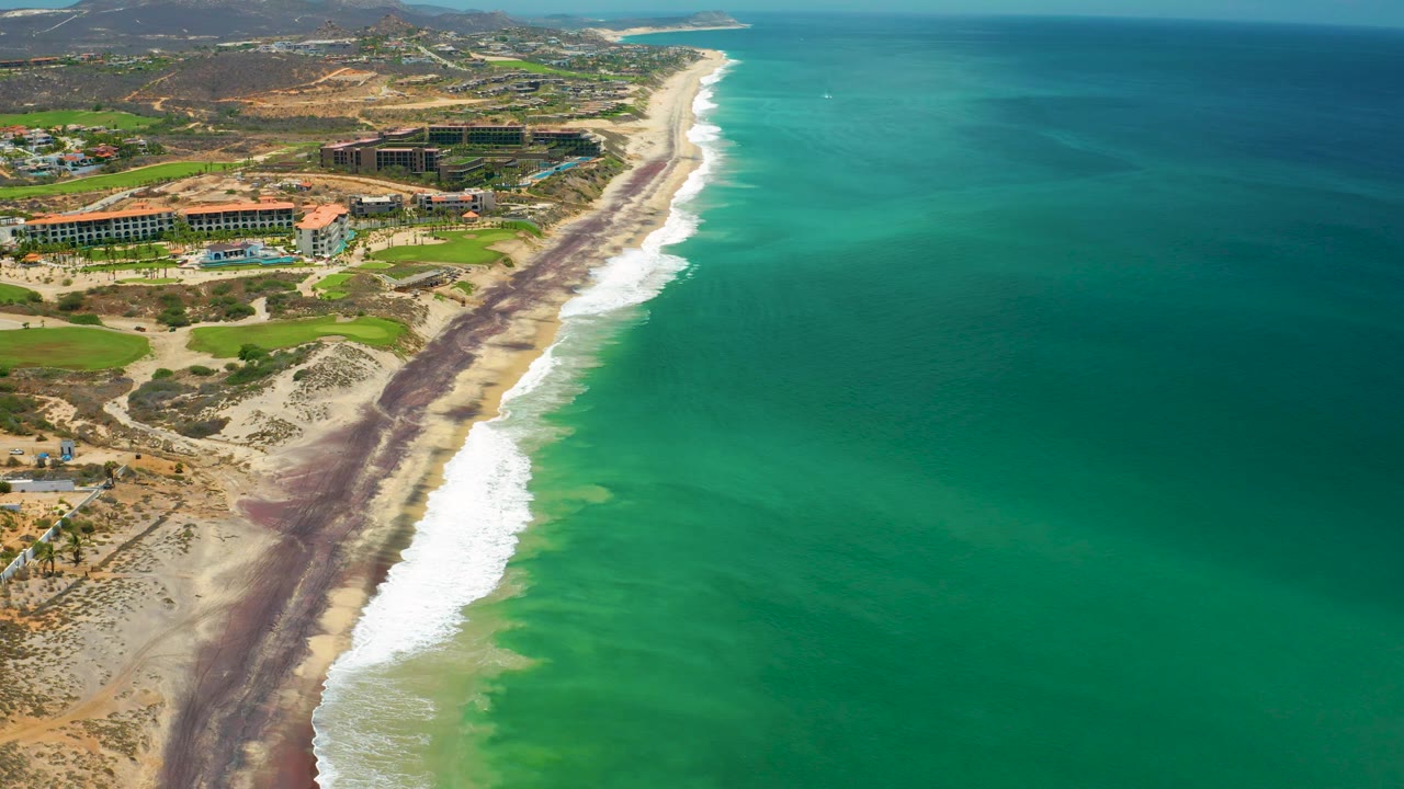San Jose del Cabo, Mexico aerial frame showing white surf breaking along a pale beach, dark bands near the waterline, golf-green landscaping and resort structures inland, and no visible people.