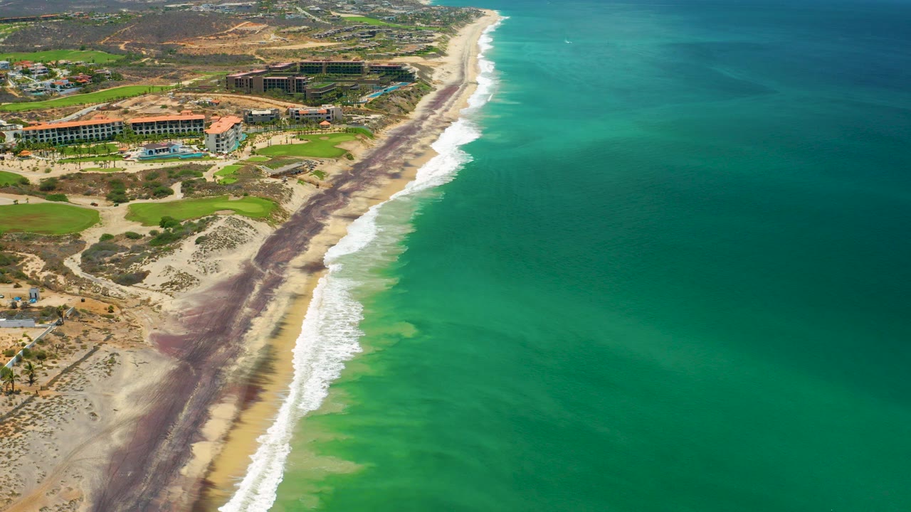 Opening aerial view in San Jose Del Cabo, Mexico, looking along green sea water and white surf beside a sandy beach with resort buildings inland and no visible people.