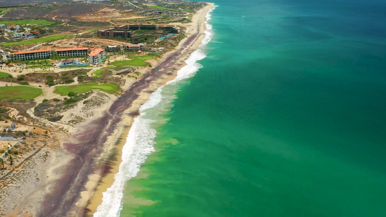 San Jose Del Cabo aerial coastline frame with a broad field of green ocean, white surf meeting sand, dark streaks along the beach, inland resort buildings, and no visible people.