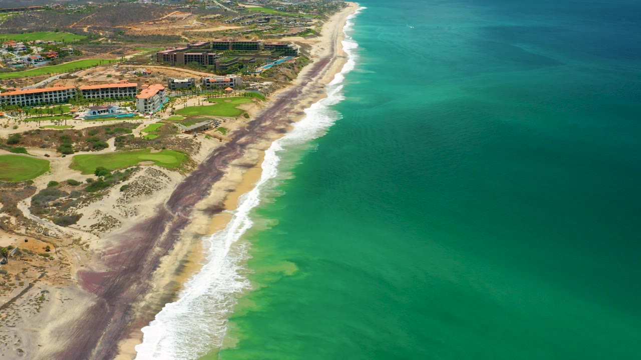 Aerial view in San Jose Del Cabo, Mexico, showing resort buildings beside a sandy beach, white surf, turquoise ocean, dry inland terrain, and no visible people.