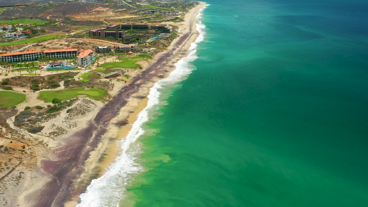 Aerial view in San Jose Del Cabo, Mexico, with more open turquoise water on the right, white wave lines along the sand, resort roofs inland, and no visible people.