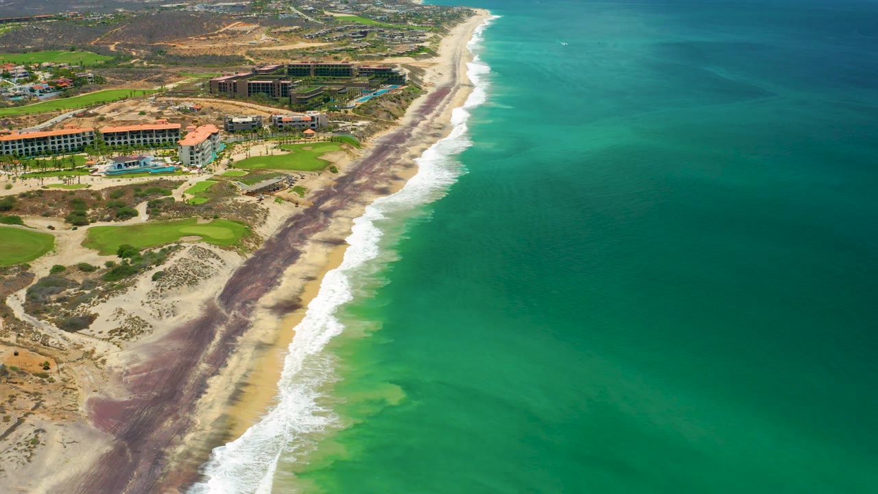 Aerial frame in San Jose Del Cabo, Mexico, showing resort grounds at left, a pale beach with dark bands near the waterline, breaking surf, green ocean, and no visible people.