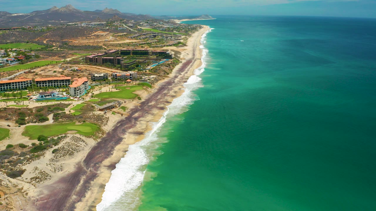 Opening aerial view over San Jose Del Cabo, Mexico, with turquoise ocean on the right, white surf along the sandy shore, resort buildings inland, mountains in the distance, and no visible people.