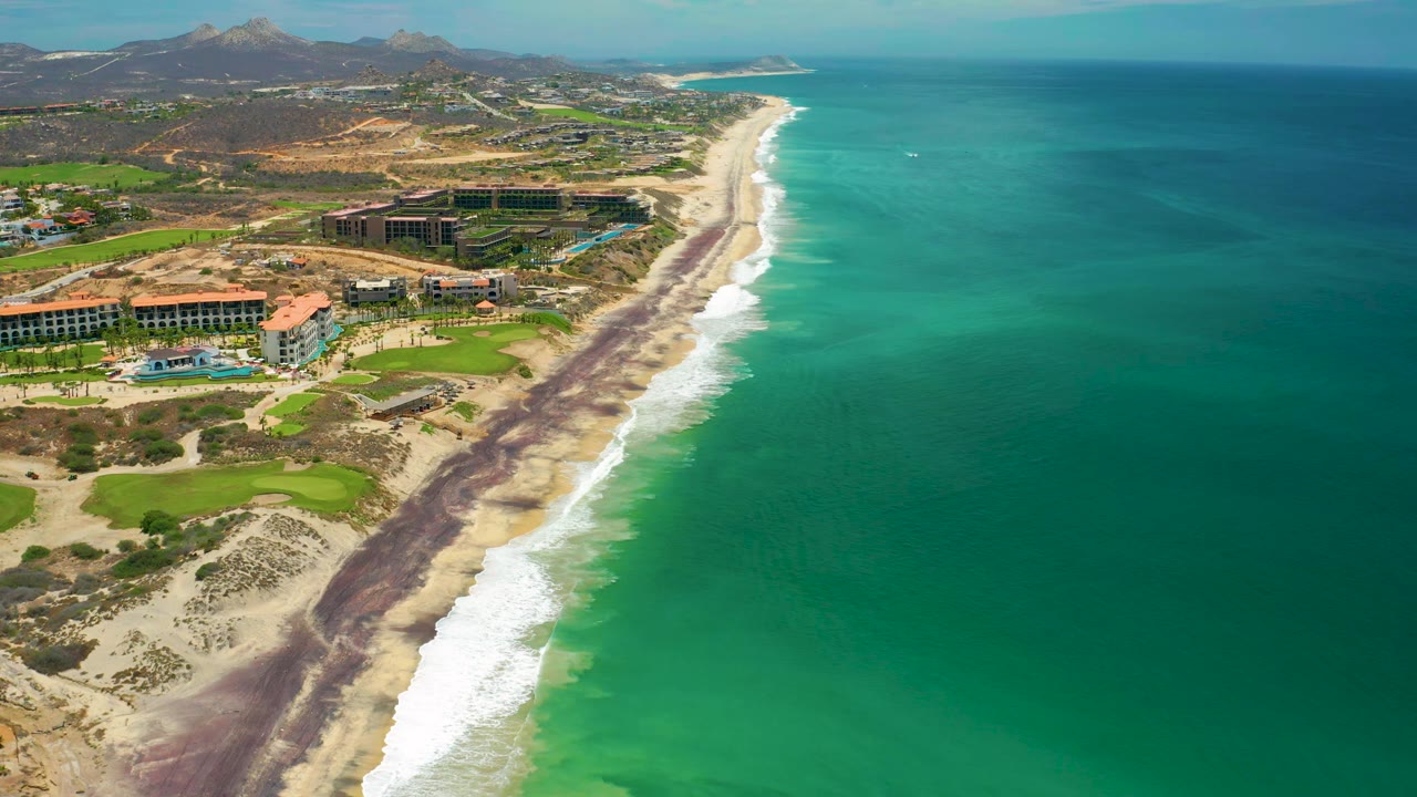Aerial frame over San Jose Del Cabo, Mexico, showing a long diagonal beach, bright blue-green water, foamy surf, resort structures, green lawns, distant mountains, and no visible people.