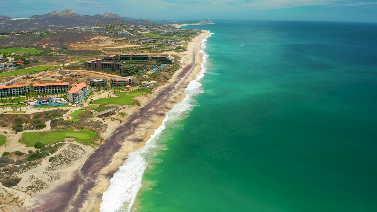 San Jose Del Cabo aerial view with more open ocean filling the right side, surf curving along the beach, resort buildings and dry hills inland, mountains behind, and no visible people.